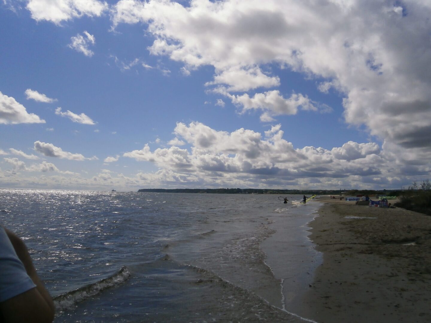 Windsurferzy na piaszczystej plaży, częściowo zachmurzone niebo i spokojne morze.