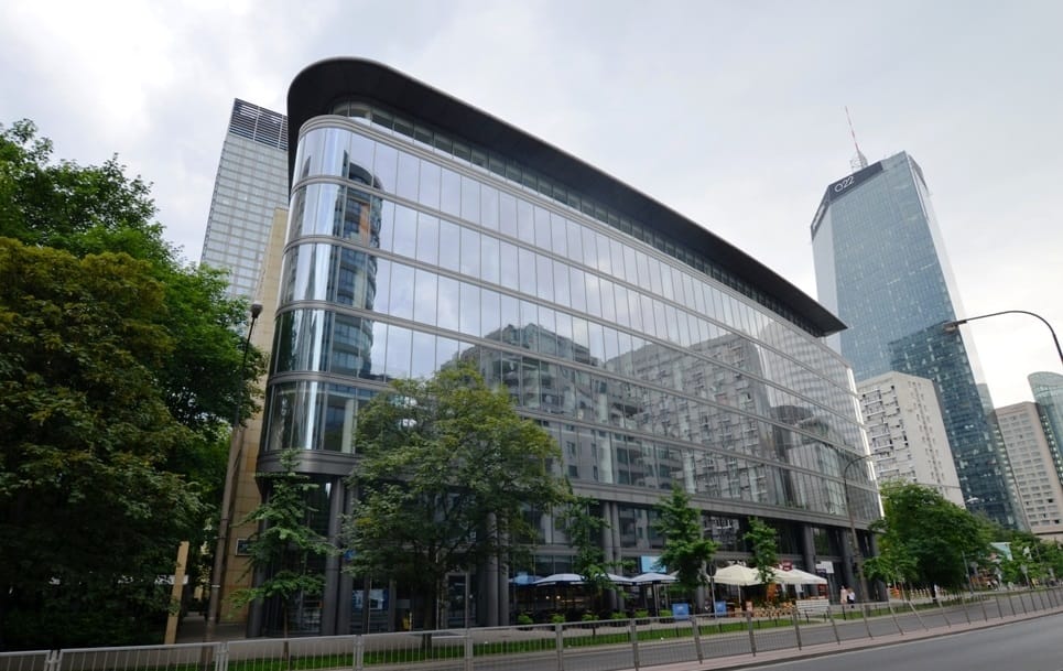 Nowoczesny szklany budynek biurowy w Warszawie, w którym odbijają się okoliczne wieżowce.