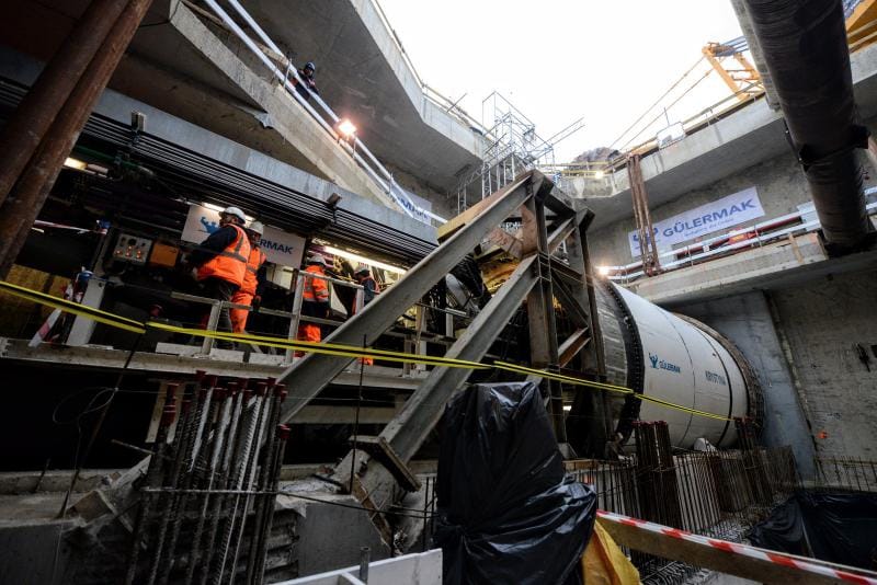 Maszyna drążąca tunele (TBM) na placu budowy, pracownicy w pomarańczowych kamizelkach bezpieczeństwa nadzorują operację. Gülermak.