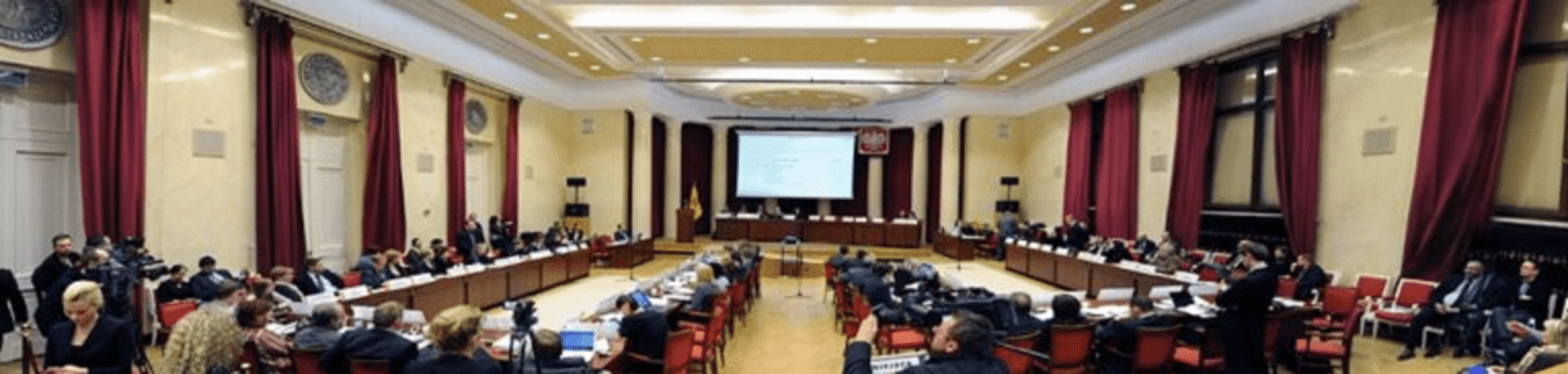 Duża sala konferencyjna, w której wiele osób siedzi przy długich stołach, uczestnicząc w spotkaniu lub dyskusji.