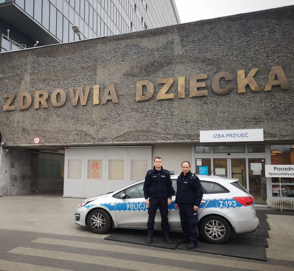 Dwóch polskich policjantów stoi przy radiowozie przed Szpitalem Zdrowia Dziecka.