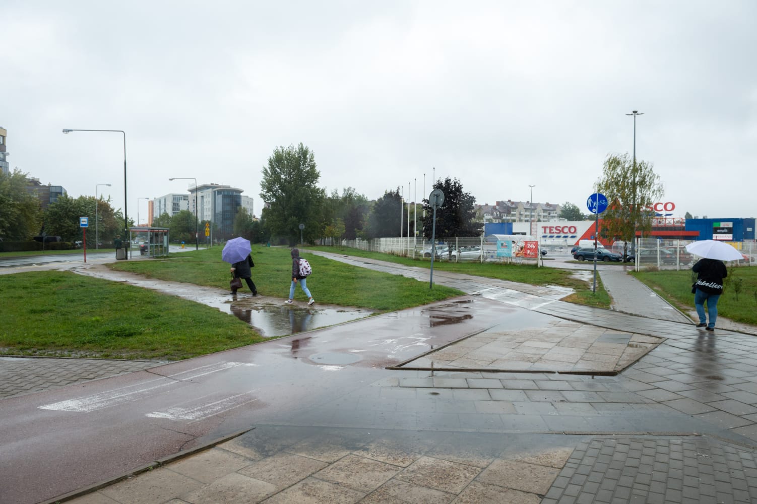 Piesi z parasolami poruszają się po zalanym deszczem skrzyżowaniu w pobliżu Tesco.