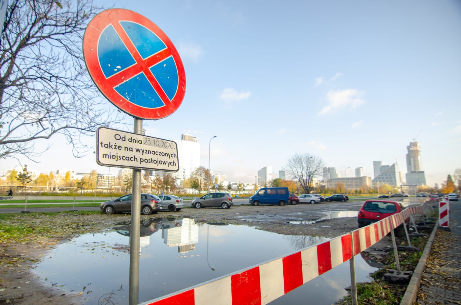 Znak zakazu parkowania w języku polskim przedstawiający samochody zaparkowane na błotnistym parkingu, w tle panorama miasta.