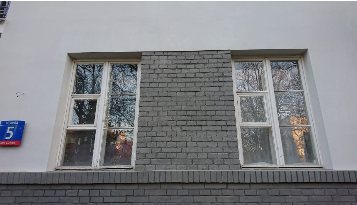 Szara, ceglana fasada budynku z dwoma wielopanelowymi oknami, w których odbijają się drzewa.