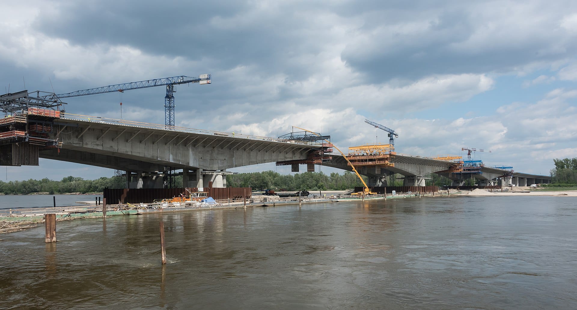 Budowa mostu nad rzeką. Widoczne są liczne dźwigi i pracownicy.