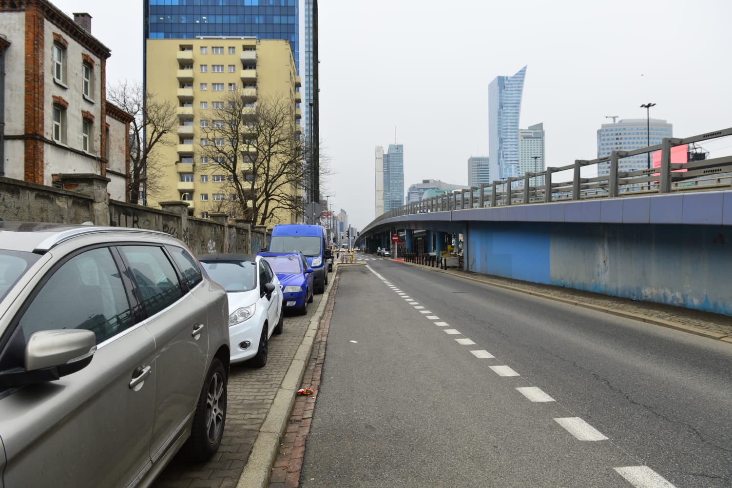 Samochody zaparkowane wzdłuż miejskiej ulicy, w tle nowoczesne wieżowce.