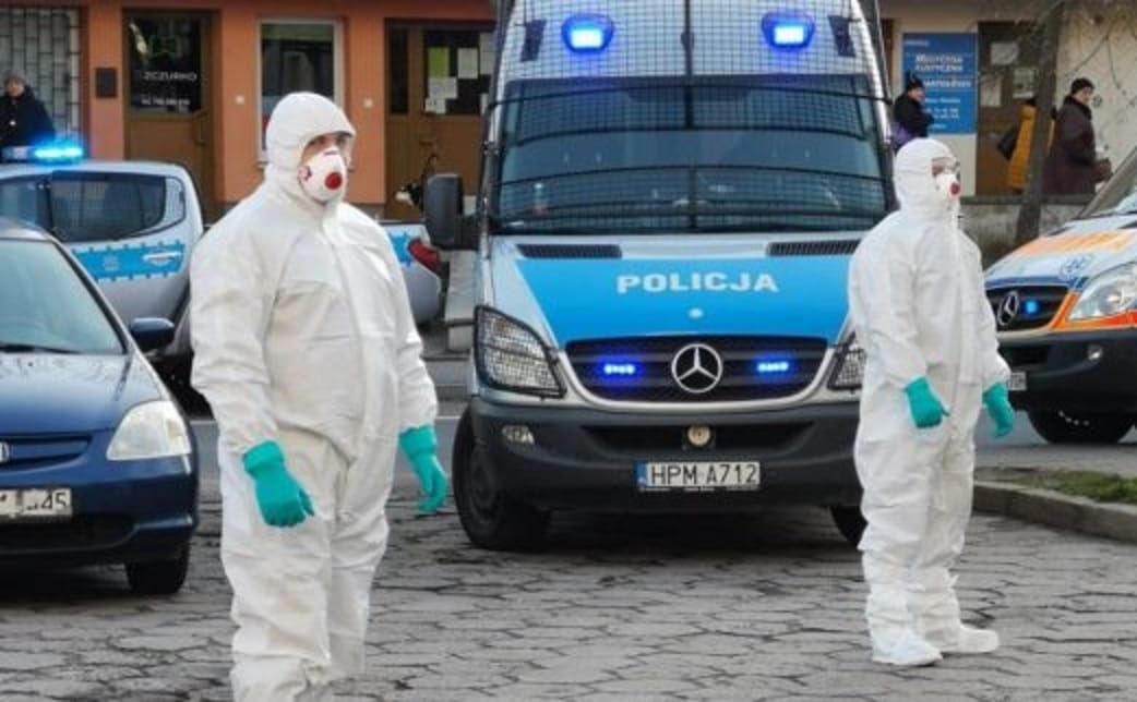 Dwóch policjantów w kombinezonach ochronnych stoi przy policyjnym furgonie. Na furgonie widoczna jest POLICJA.