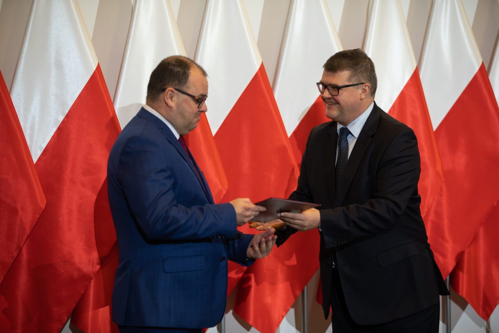 Dwóch mężczyzn w garniturach wymienia dokumenty przed polskimi flagami.