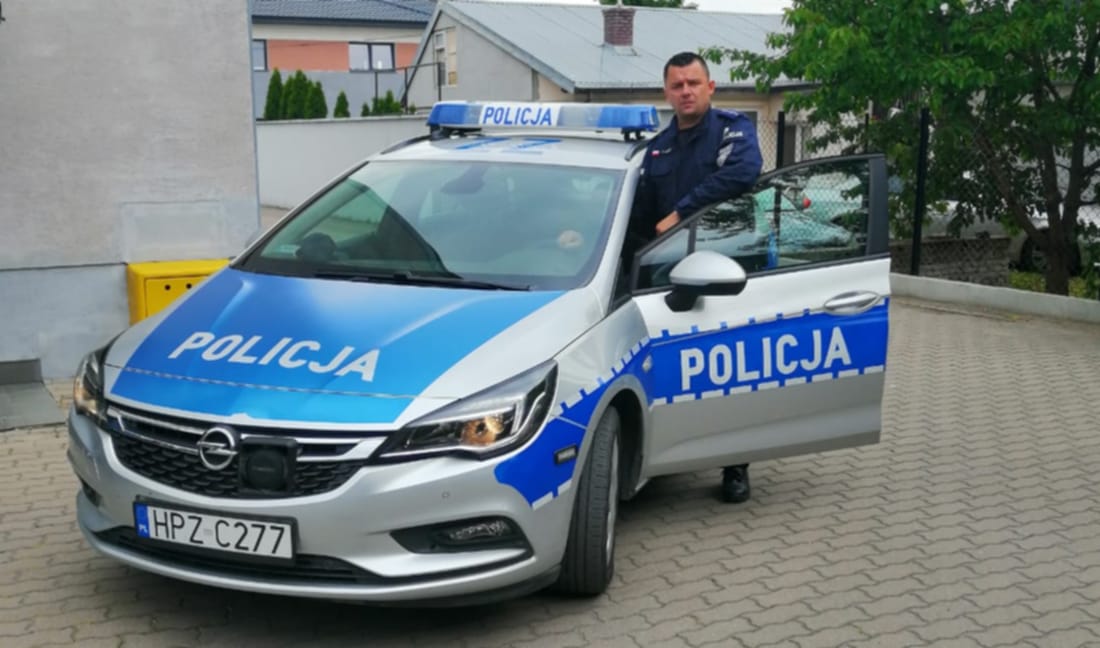 Polski policjant stoi obok srebrnego radiowozu Opel Astra. Na tablicy rejestracyjnej samochodu widnieje HPZ C277.