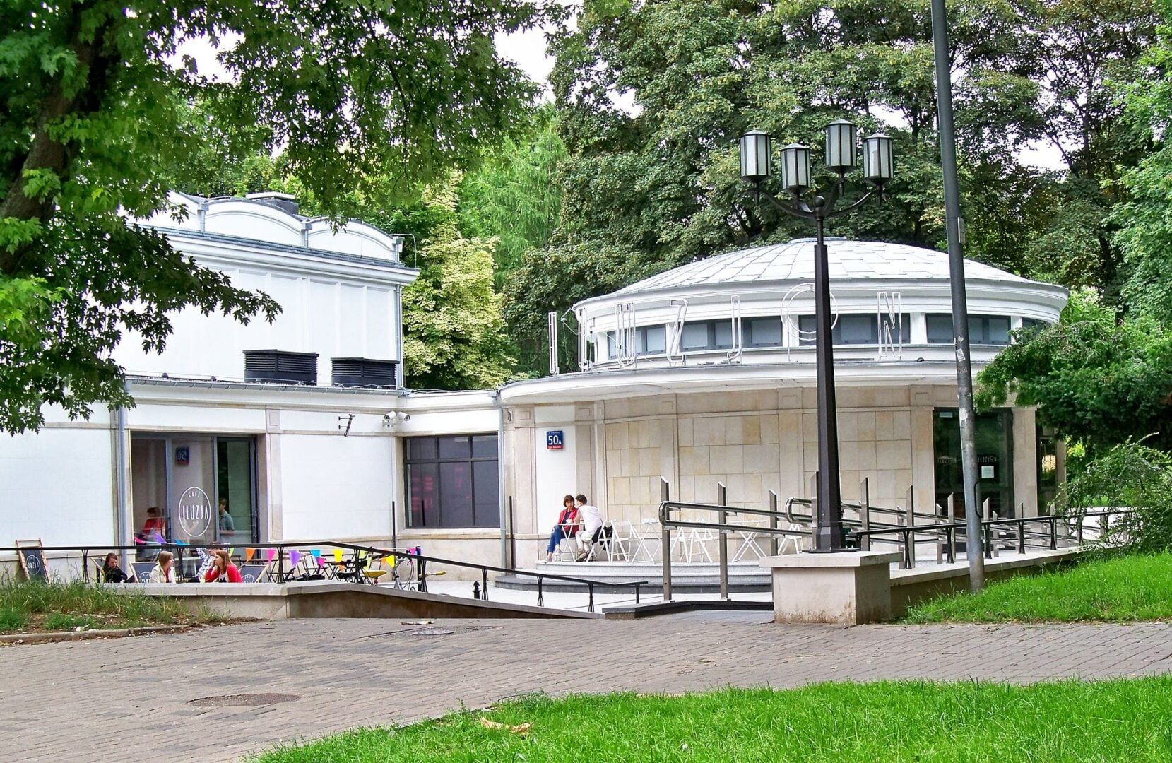 Taras na świeżym powietrzu Cafe Illuzin w warszawskim parku. Goście odpoczywają przy stolikach pod drzewami.