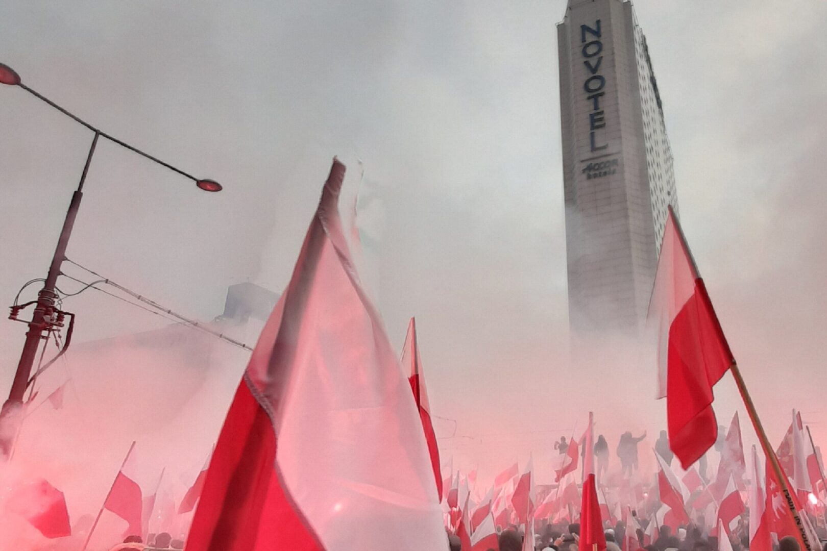 Tłum ludzi machających polskimi flagami pośród czerwonego dymu, w tle hotel Novotel.