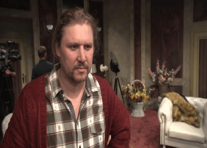 Mężczyzna z długimi włosami, ubrany w czerwony kardigan i kraciastą koszulę, udziela wywiadu.