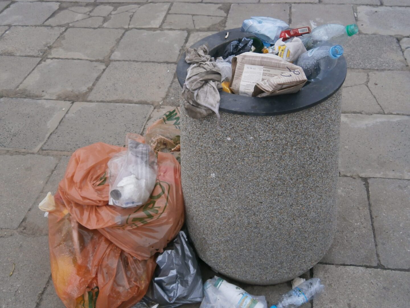 Przepełniony kosz na śmieci pełen plastikowych butelek i toreb na chodniku w mieście.