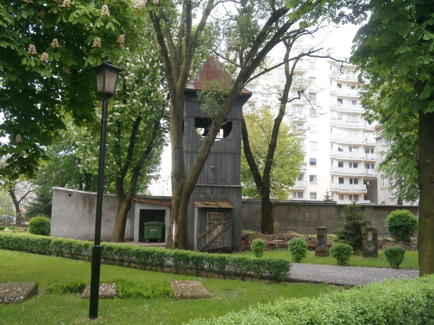 Stara drewniana dzwonnica na cmentarzu, otoczona drzewami i nagrobkami.