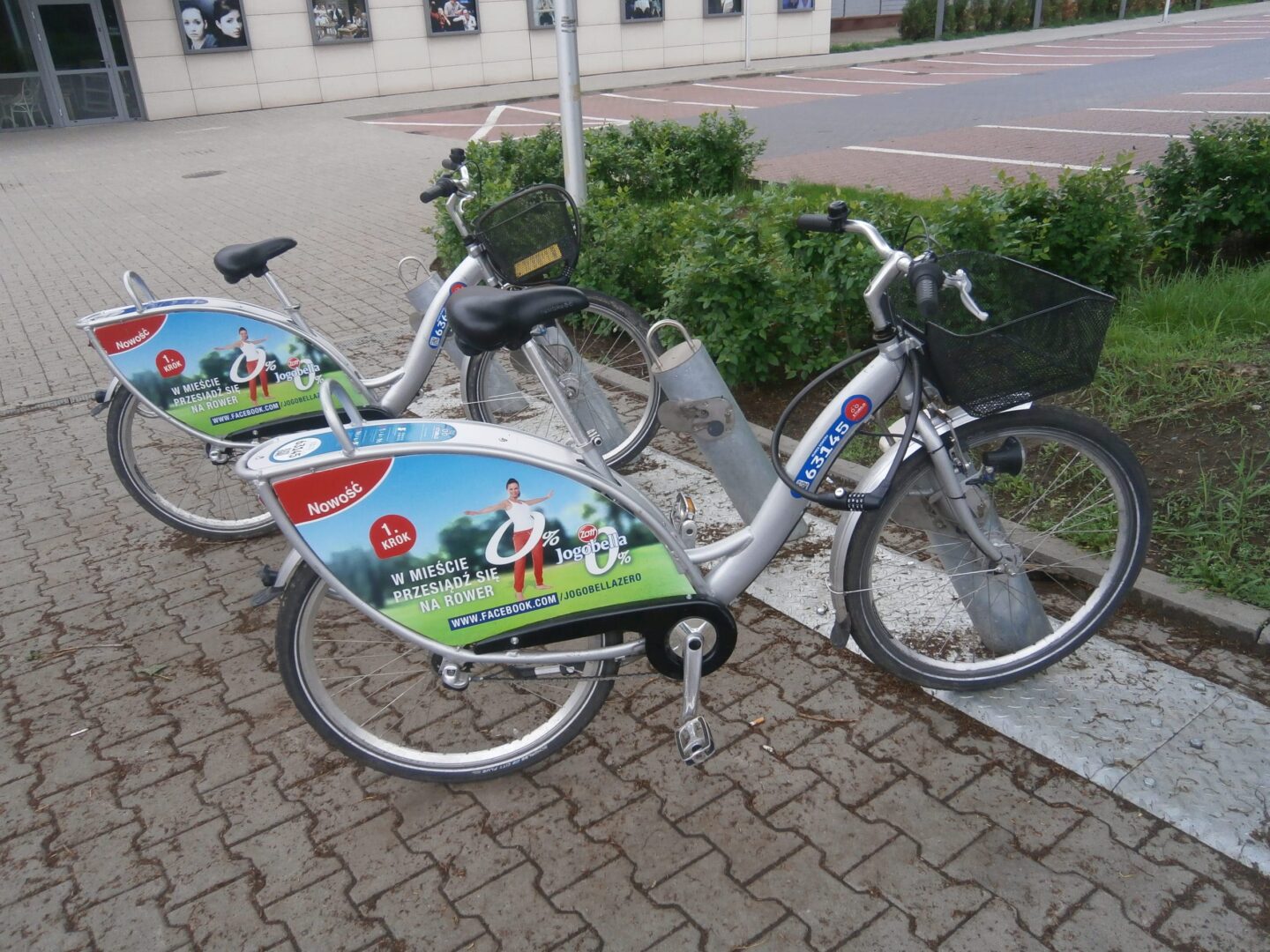 Dwa srebrne rowery miejskie z reklamą Jogobella zaparkowane na ceglanym chodniku.