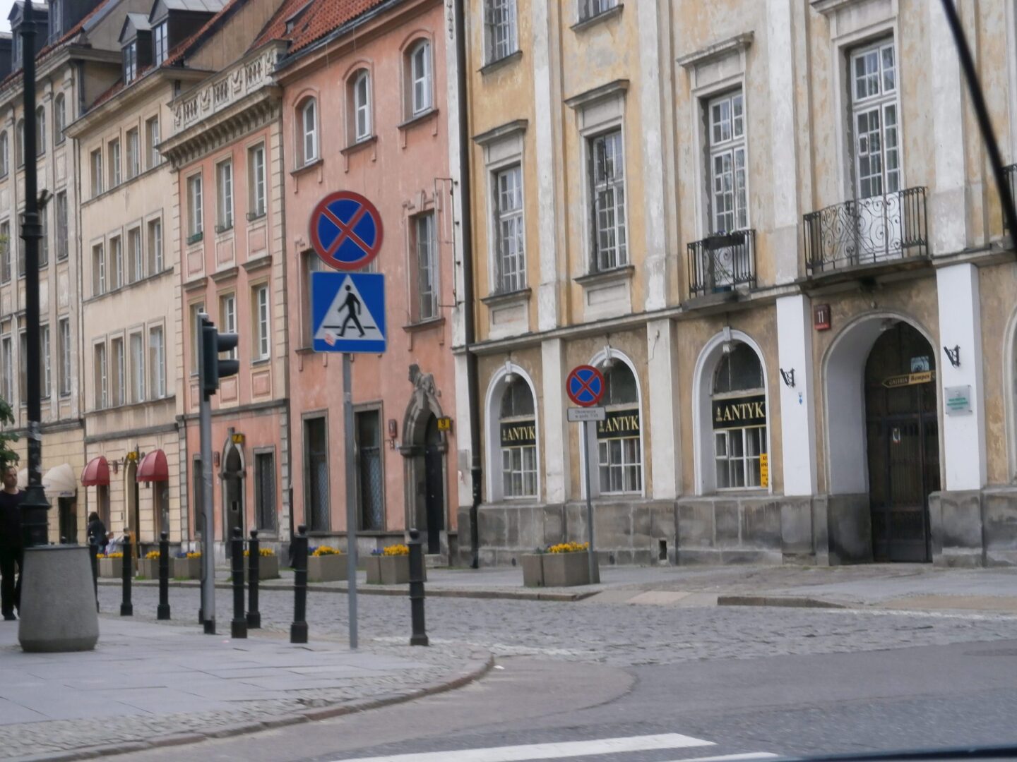 Widok na ulicę Warszawy z zabytkowymi budynkami, przejściem dla pieszych i znakami zakazu parkowania.