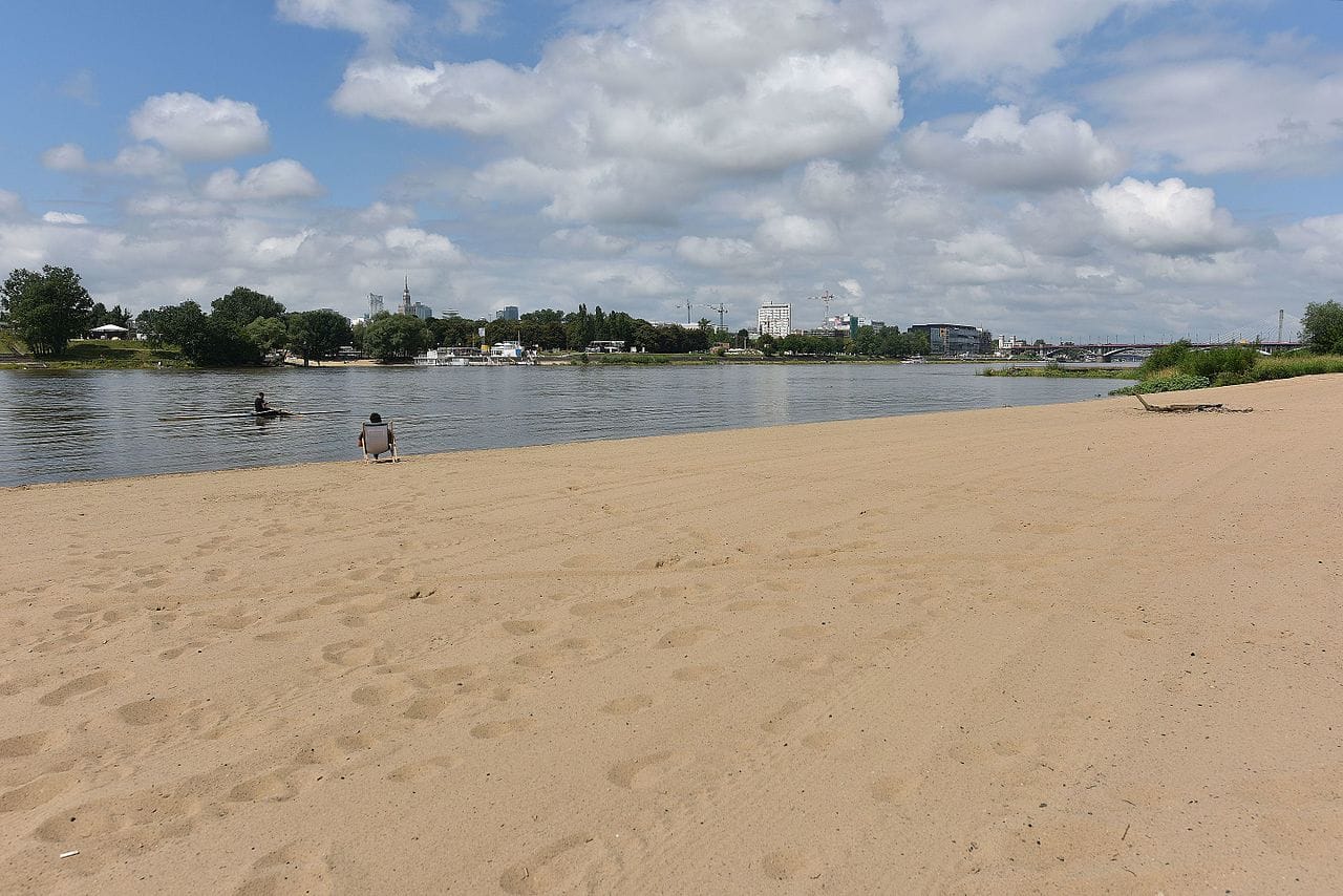 Osoba odpoczywająca na leżaku na piaszczystym brzegu rzeki, z widokiem na panoramę miasta.