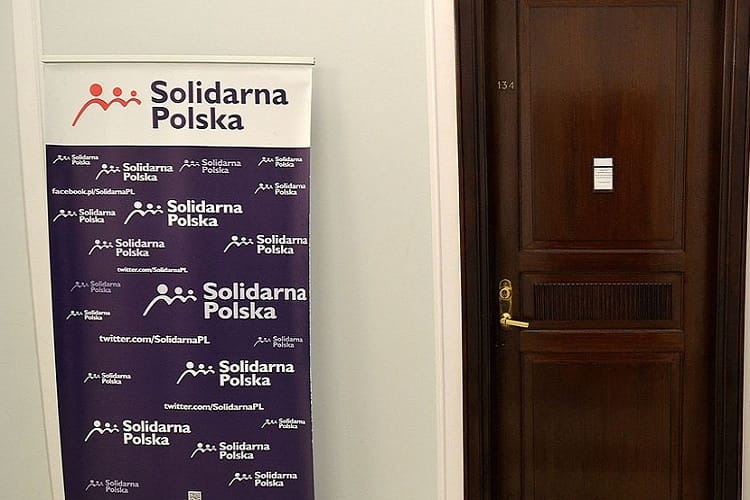 Logo Solidarnej Polski na fioletowym banerze obok ciemnobrązowych drzwi.