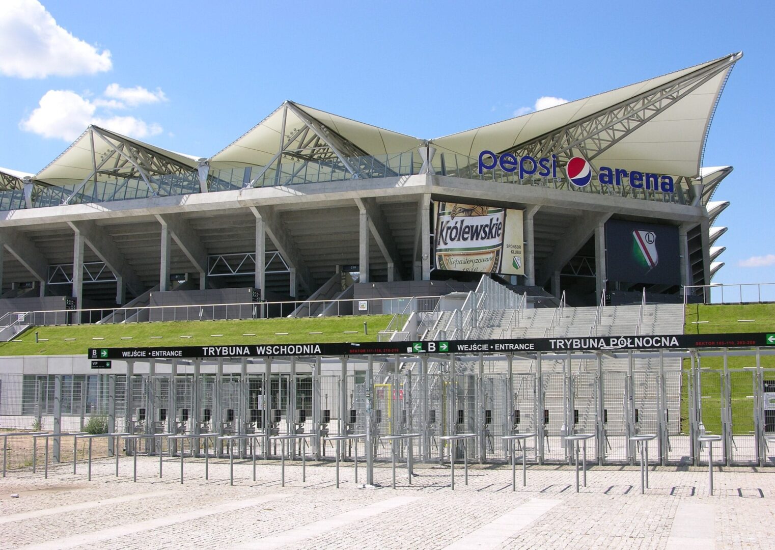 Zewnętrzna część Pepsi Areny z bramami wejściowymi i tabliczkami z napisami „Trybuna Wschodnia” i „Trybuna Północna”.