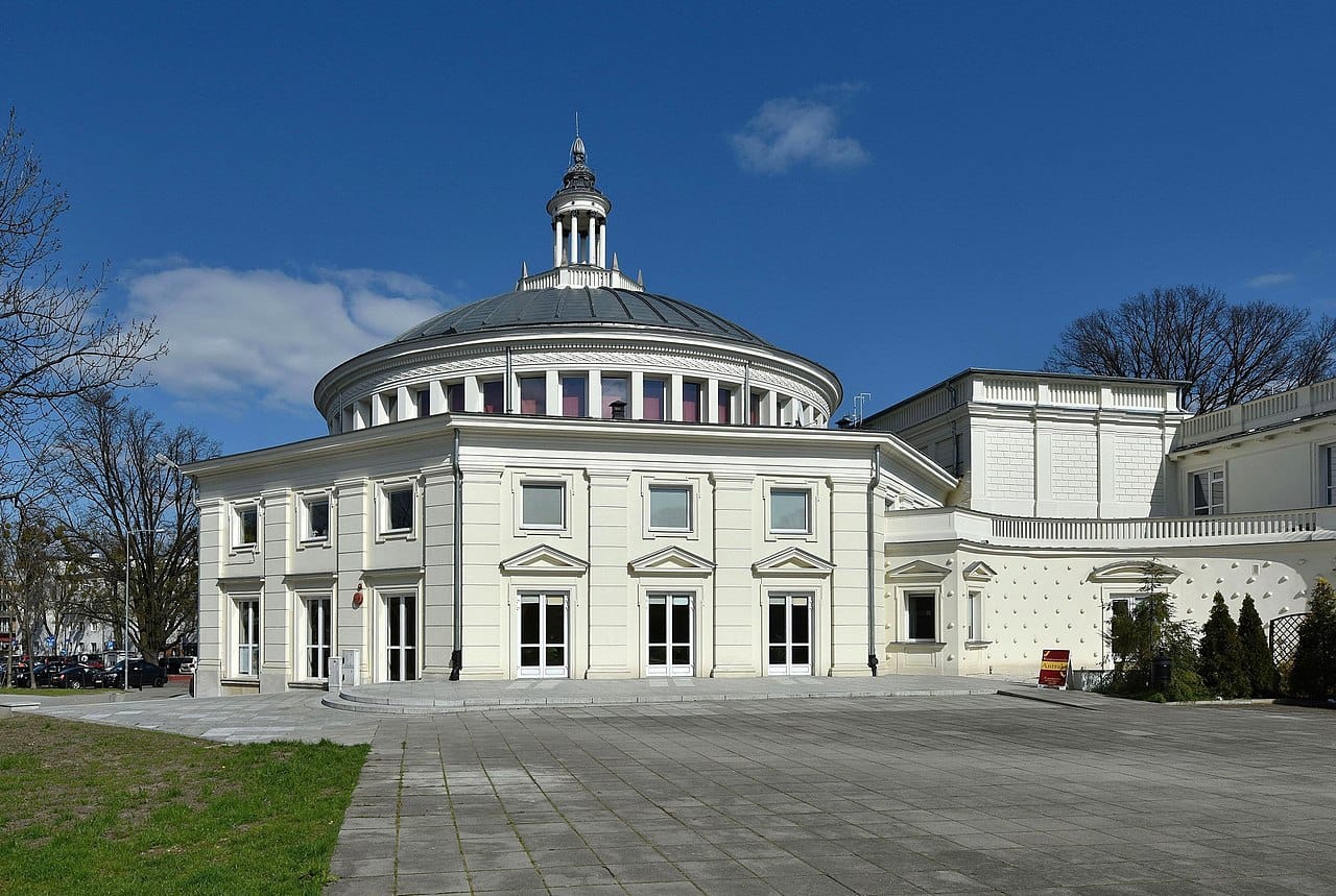 Elegancki biały budynek w kształcie rotundy z kopułą, otoczony brukowanym placem.