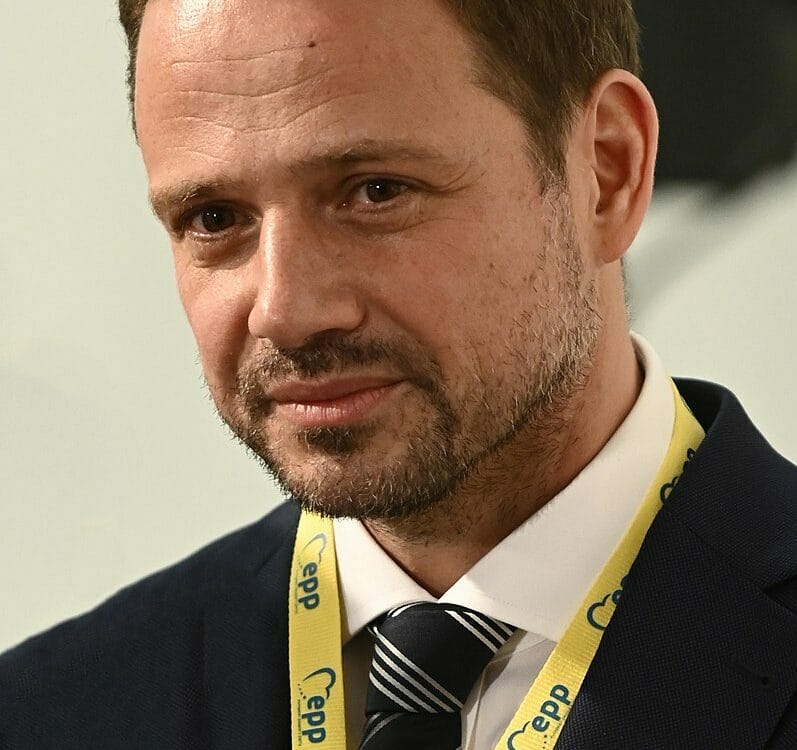 Portret mężczyzny z krótkimi brązowymi włosami i brodą, ubranego w ciemny garnitur i krawat w paski. Ma na sobie smycz z napisem „Cepp”.