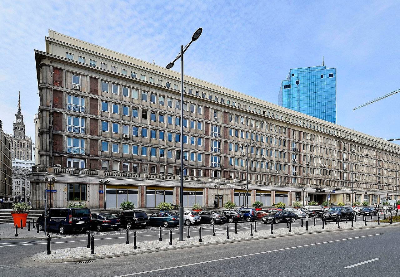 Modernistyczna architektura Warszawy: długi, niski budynek z wieloma oknami, zaparkowane przed nim samochody i nowoczesny wieżowiec w tle.