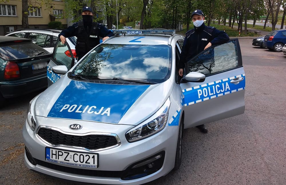 Dwóch polskich policjantów w maskach na twarzach dumnie stoi obok policyjnego samochodu Kia Ceed. Na tablicy rejestracyjnej samochodu widnieje HPZ C021.