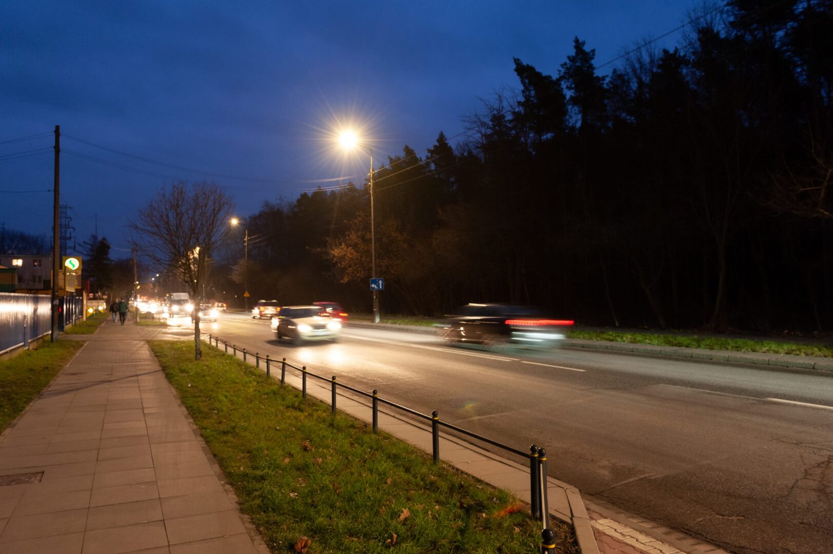 Nocna scena uliczna z samochodami przejeżdżającymi obok chodnika i drzew.