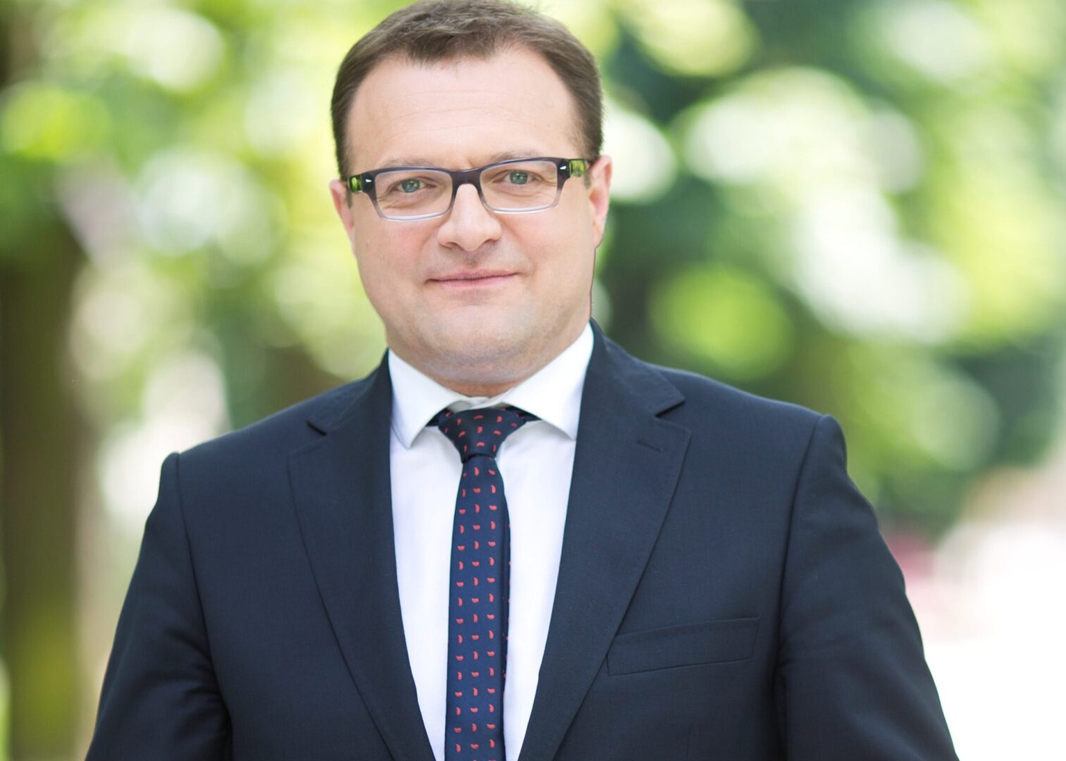 Portret mężczyzny w ciemnym garniturze i czerwono-granatowym krawacie.