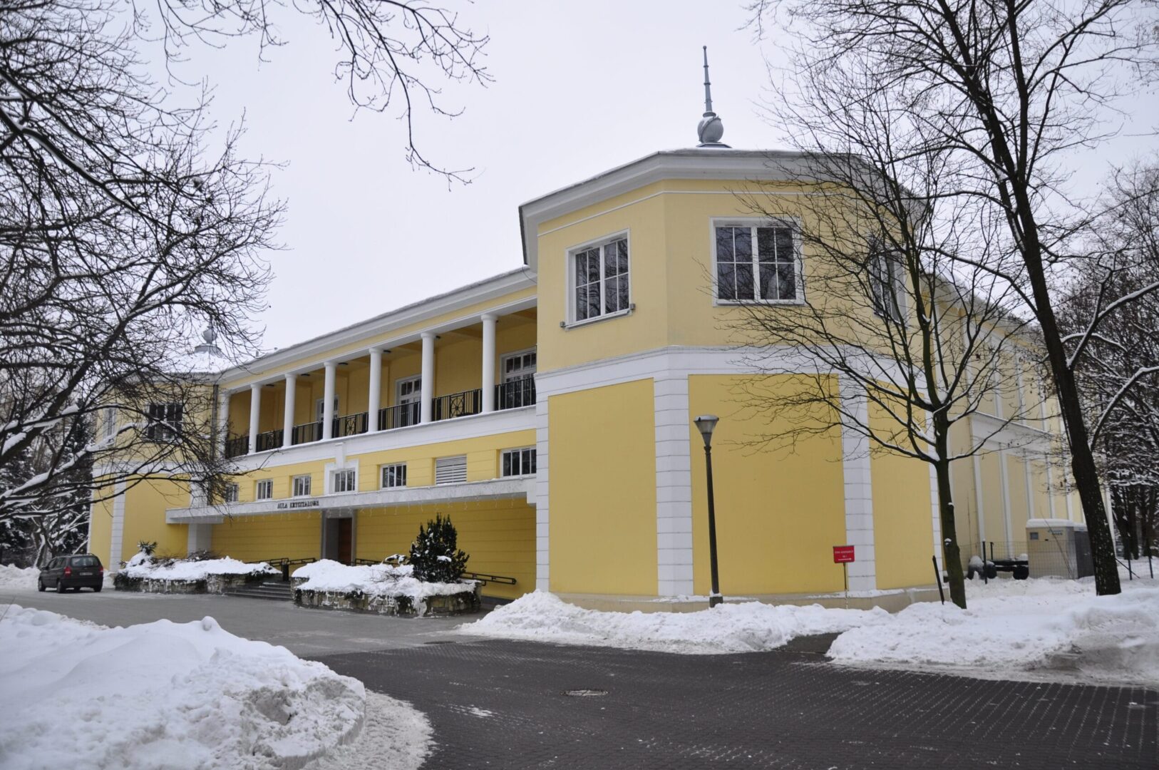 Zimowy widok Auli Uniwersyteckiej – żółtego budynku z kolumnami i pokrytym śniegiem terenem.