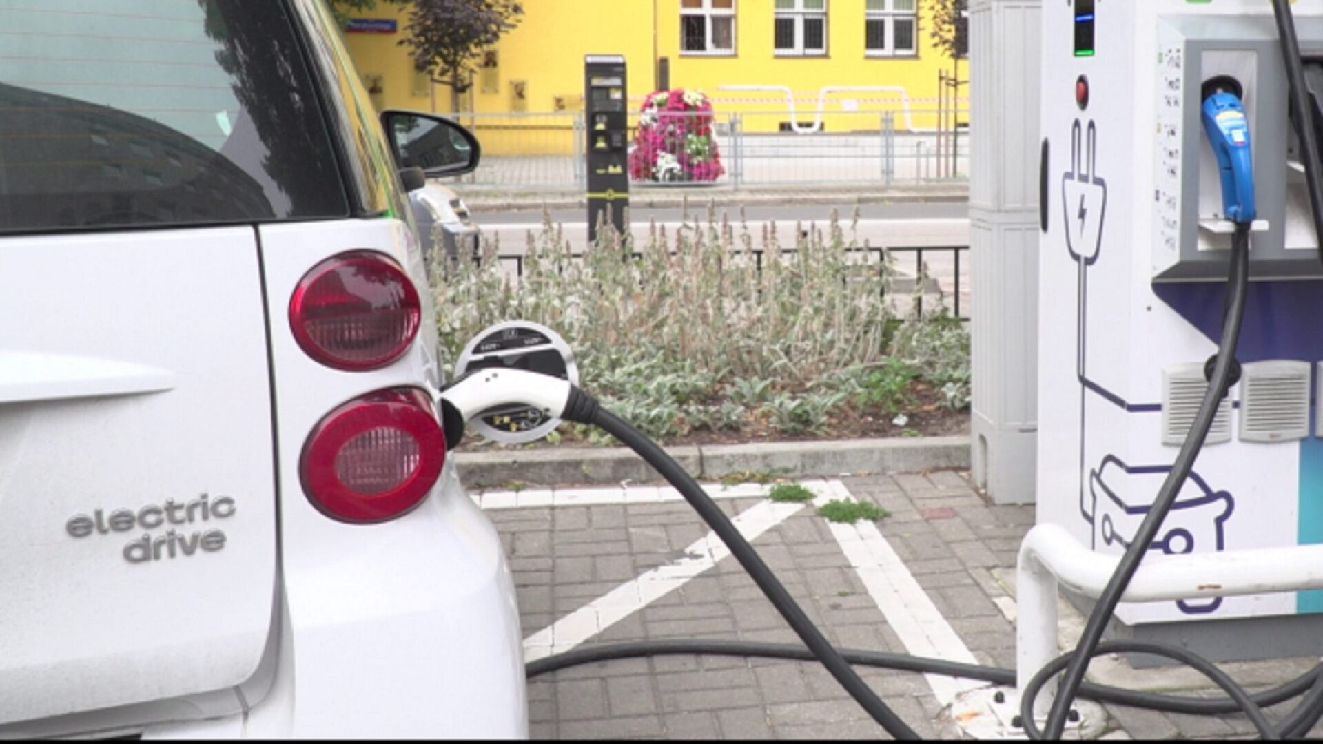 Biały samochód elektryczny ładuje się na publicznej stacji ładowania. Na samochodzie widoczny jest napis „Electric drive”.
