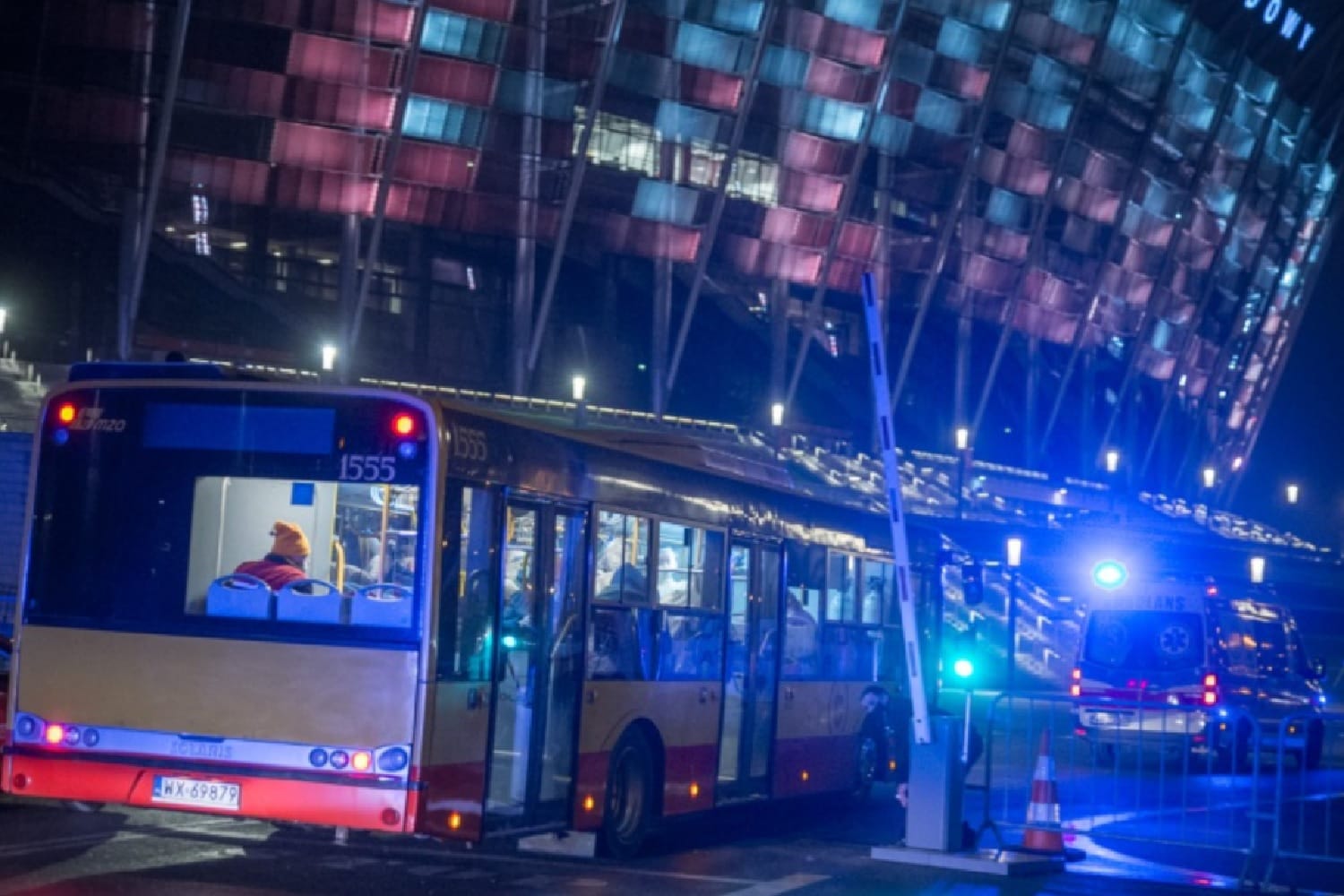 Nocna scena autobusu miejskiego zatrzymanego w pobliżu stadionu z obecnymi pojazdami ratunkowymi. Widoczny autobus numer 1555.