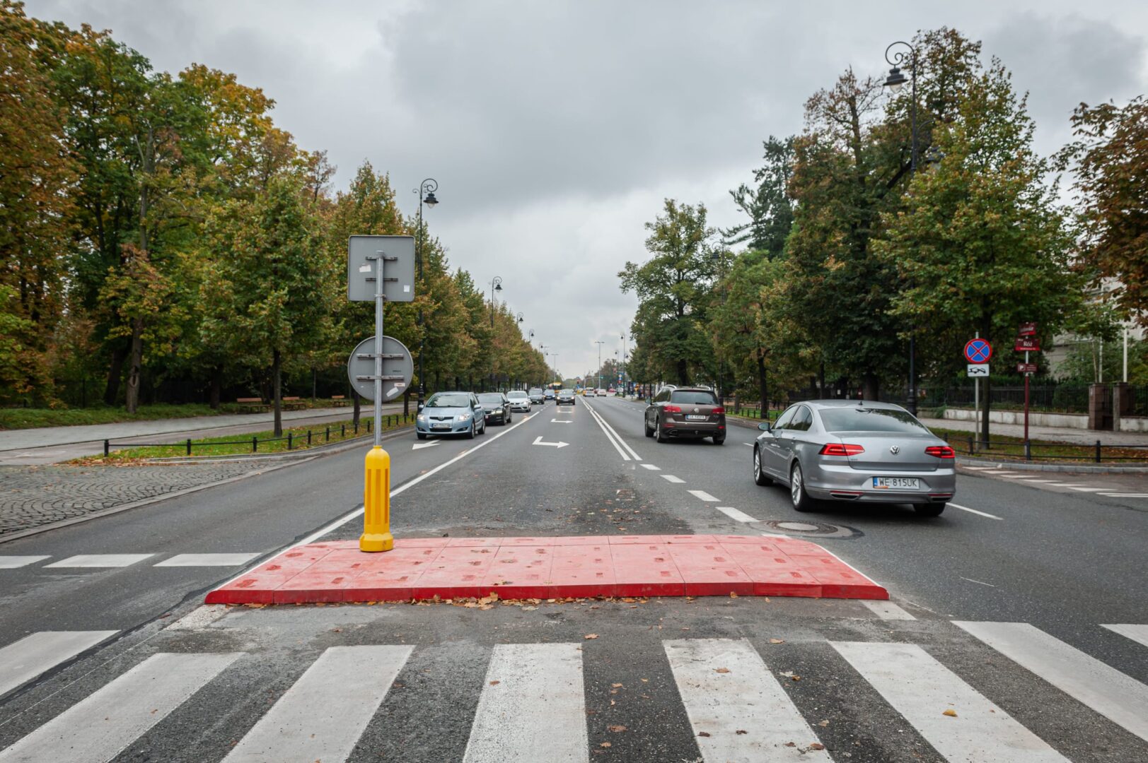 Przejście dla pieszych z czerwoną, widoczną nawierzchnią i samochodami na ulicy obsadzonej drzewami.