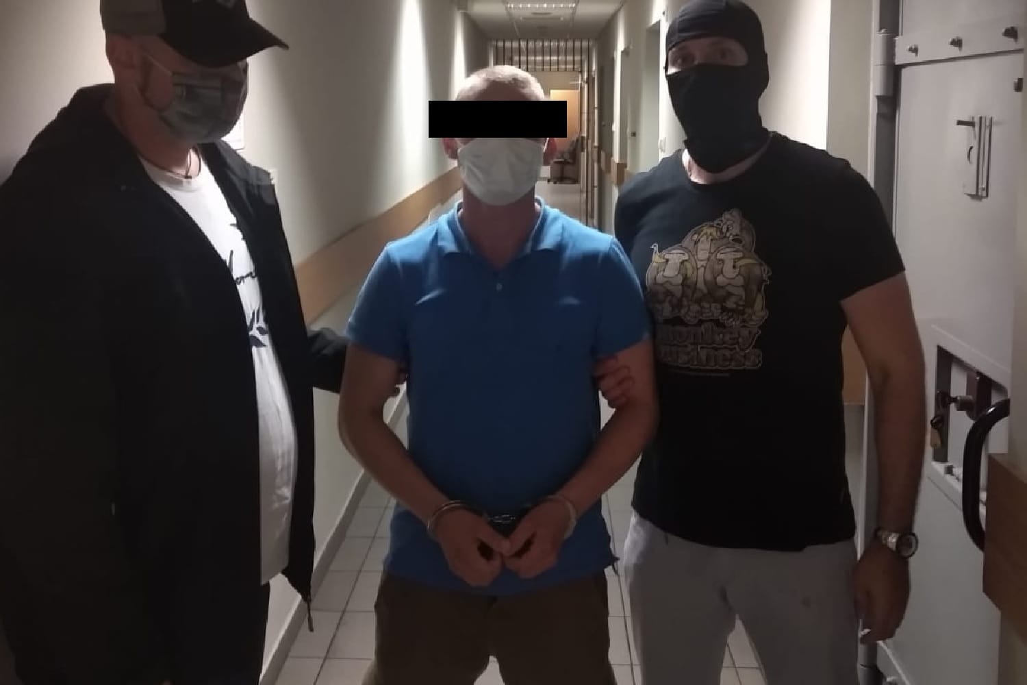 Skutego kajdankami mężczyznę eskortowanego przez dwóch funkcjonariuszy korytarzem więziennym.