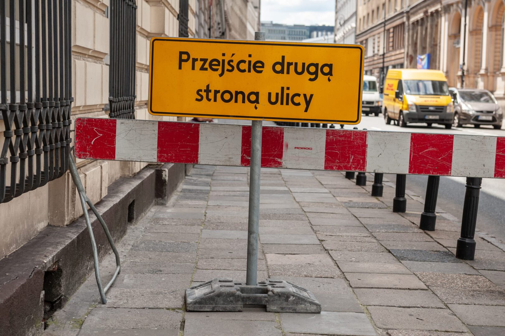 Znak informujący o zamknięciu drogi w języku polskim: „Przejście drugą stroną ulicy” z czerwonymi i białymi barierkami.