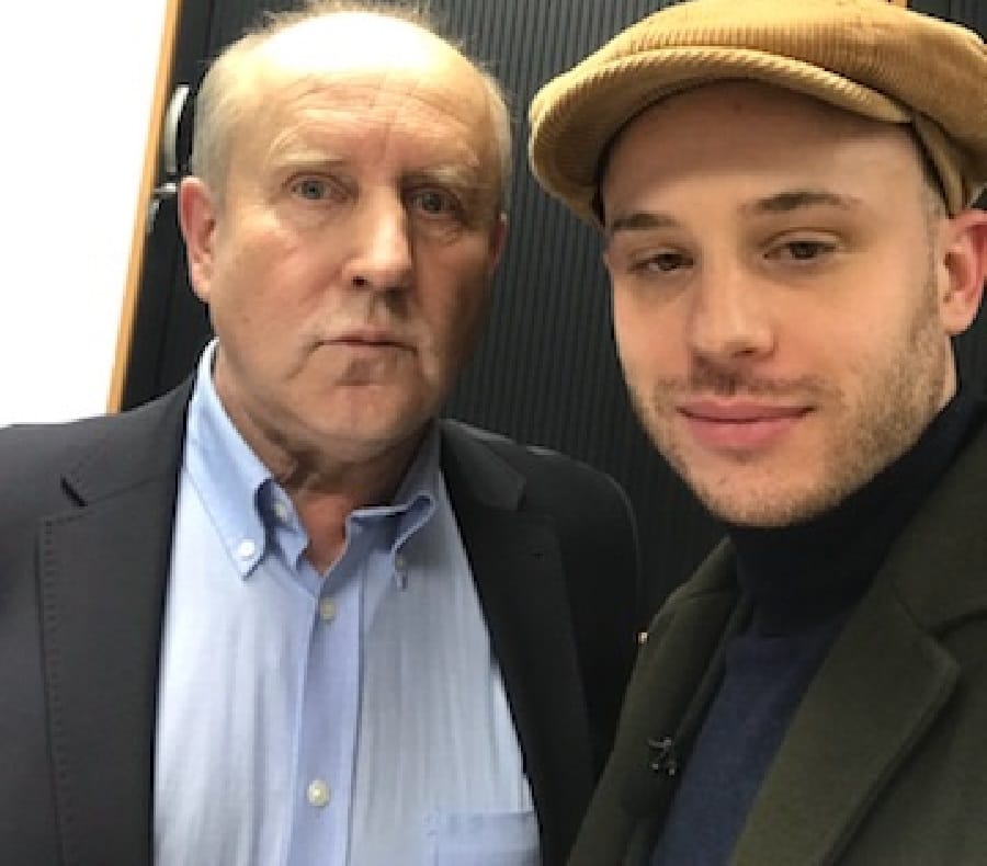 Dwóch mężczyzn pozuje do zdjęcia. Jeden mężczyzna ma na sobie garnitur, drugi czapkę gazeciarza.