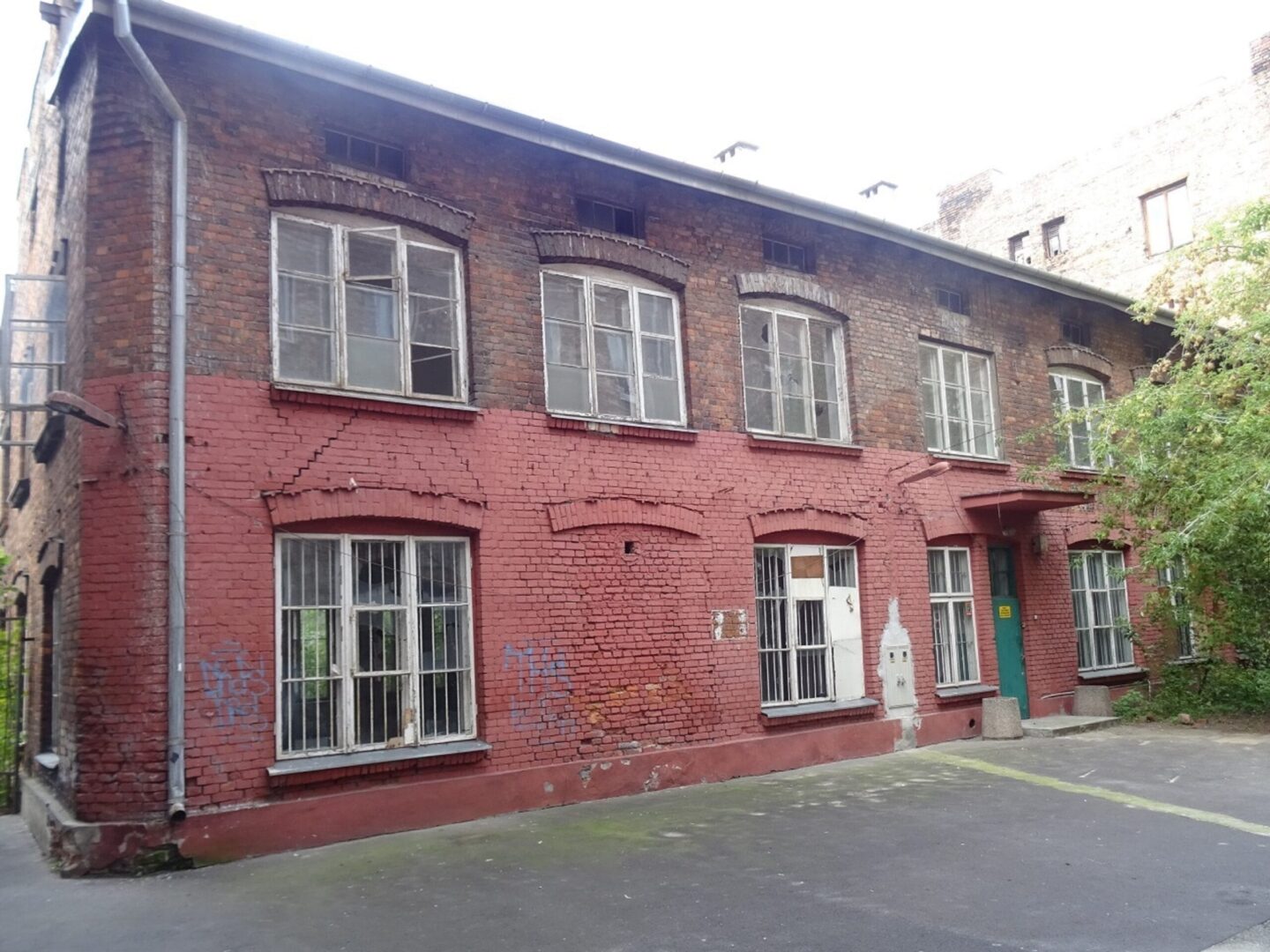 Stary ceglany budynek z łukowatymi oknami i zielonymi drzwiami. Czerwona ceglana fasada nosi ślady wieku i zużycia.