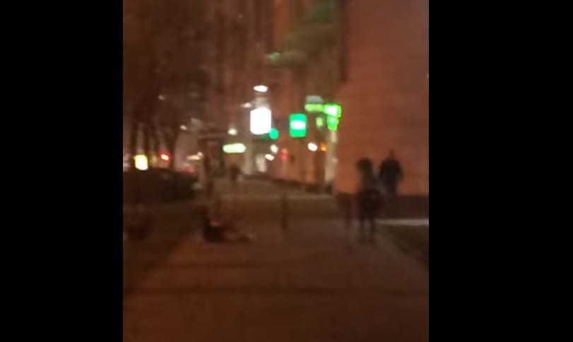 Rozmazany nocny widok osoby leżącej na ziemi, prawdopodobnie rannej, z innymi osobami w pobliżu. Incydent Grochowski.