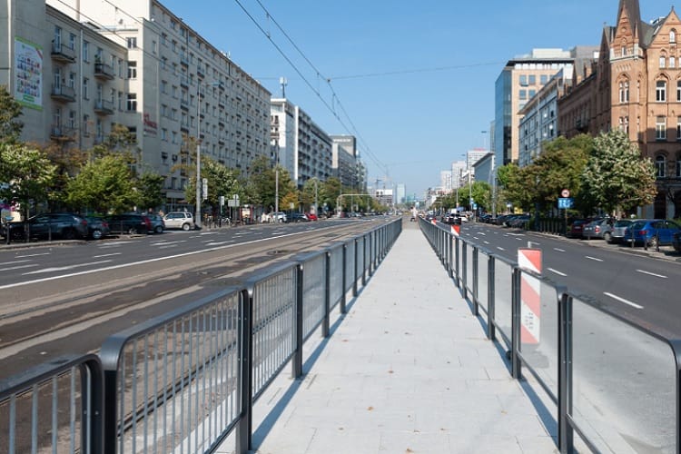 Pusta ulica miejska z torami tramwajowymi, nowoczesnymi budynkami i chodnikiem dla pieszych oddzielonym barierkami bezpieczeństwa.