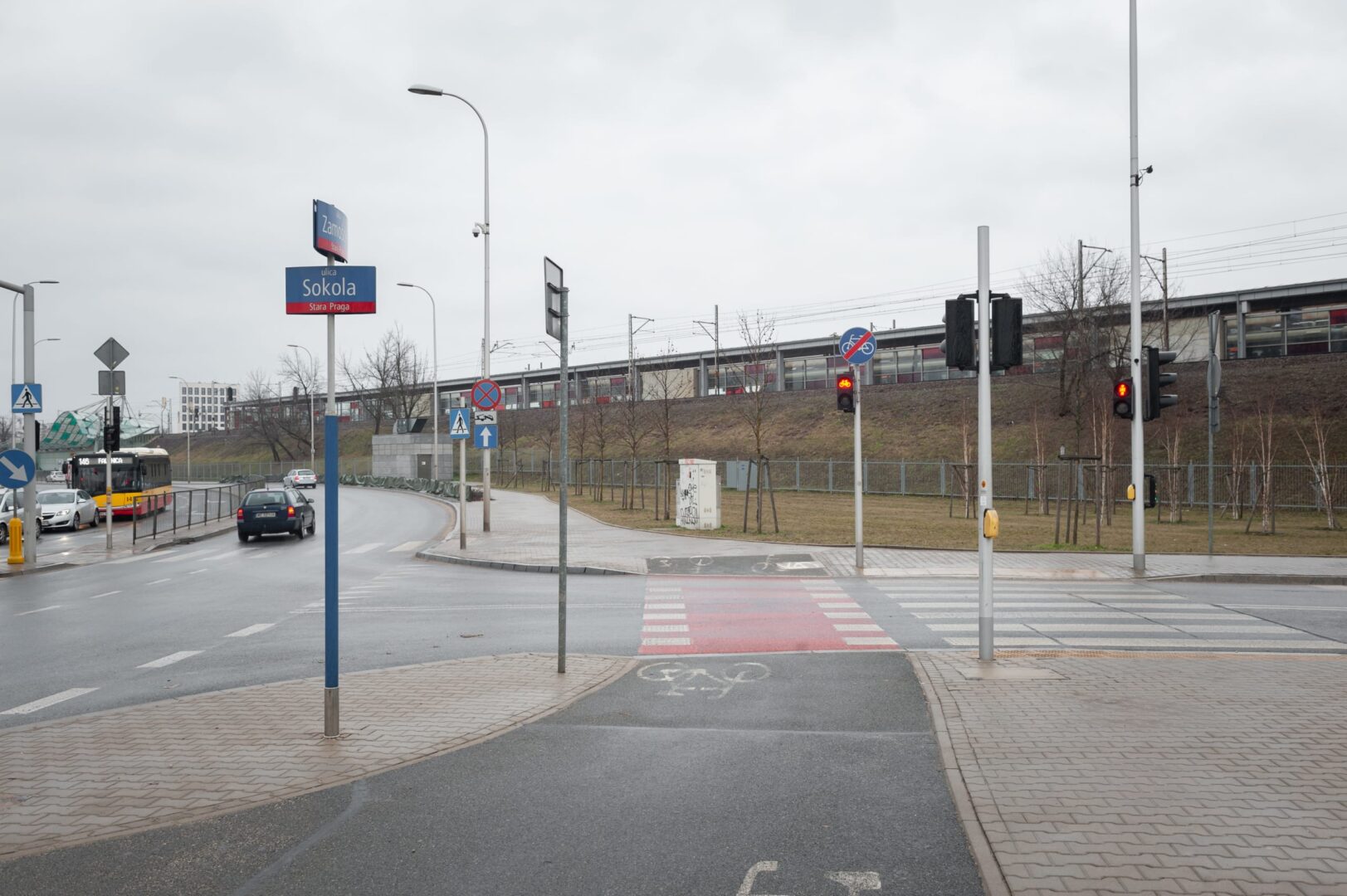 Scena uliczna w Warszawie ze znakiem ul. Sokola, przejściem dla pieszych i dworcem kolejowym w tle.