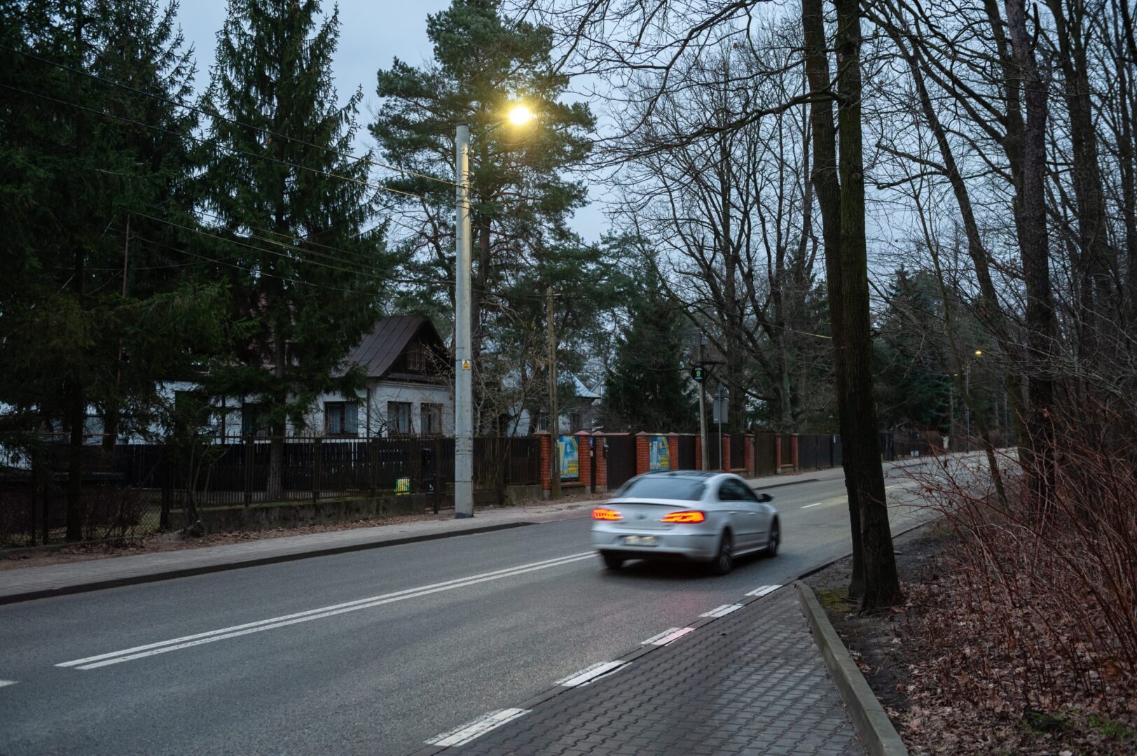 Srebrny samochód jadący ulicą osiedlową o zmierzchu, mijając domy i drzewa.