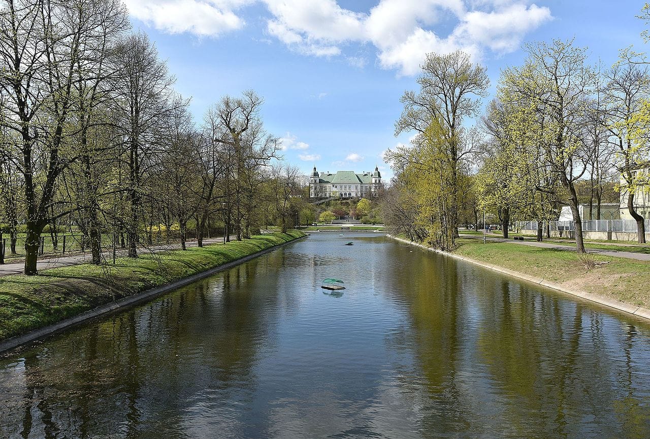 Spokojny kanał, w którego wodach odbija się pałac, brzegi porośnięte wiosennymi drzewami.