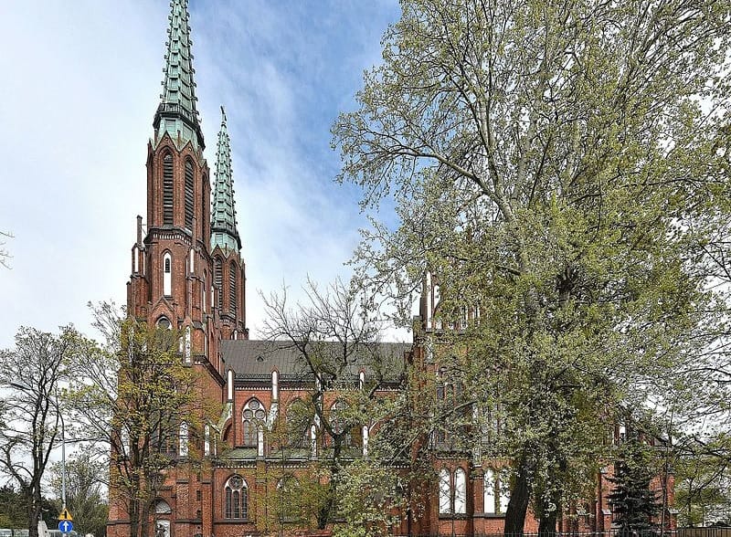 Ceglana katedra z dwiema wysokimi, ozdobnymi wieżami pod częściowo zachmurzonym niebem.