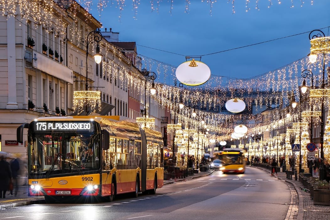 Nocna scena przedstawiająca ulicę miejską udekorowaną świątecznymi światłami, z widocznym żółtym autobusem miejskim (175 PL. Piłsudskiego 9902).
