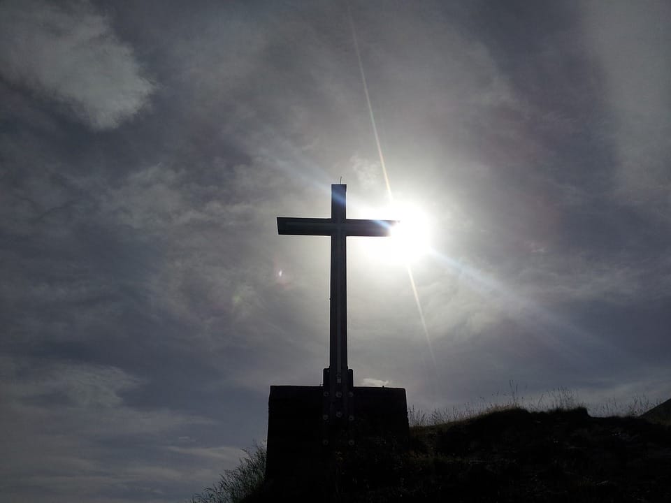 Sylwetka krzyża na szczycie wzgórza, za którym świeci słońce.