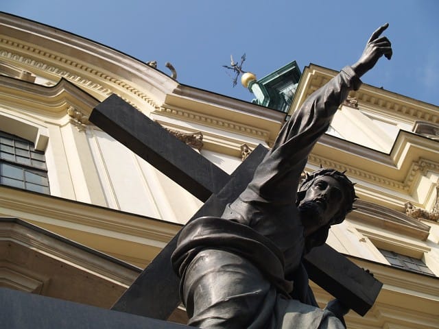 Brązowa statua Jezusa Chrystusa niosącego krzyż na tle barokowego budynku.