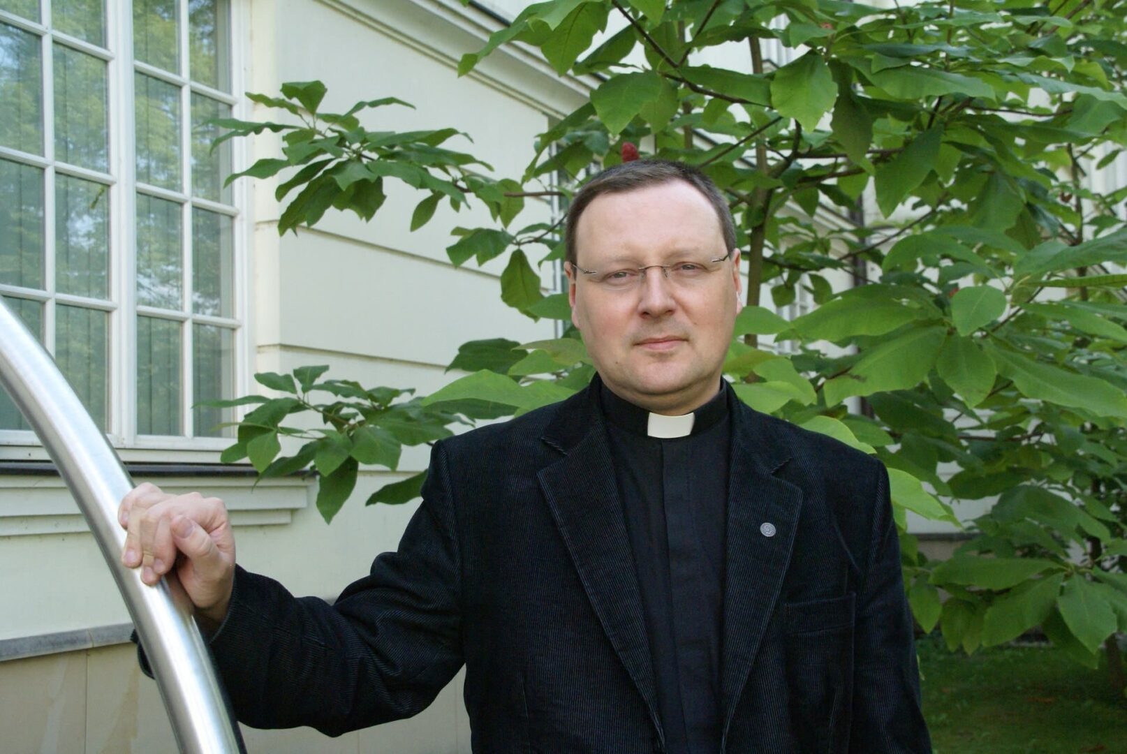 Portret księdza w czarnej kurtce, stojącego na zewnątrz w pobliżu drzewa.