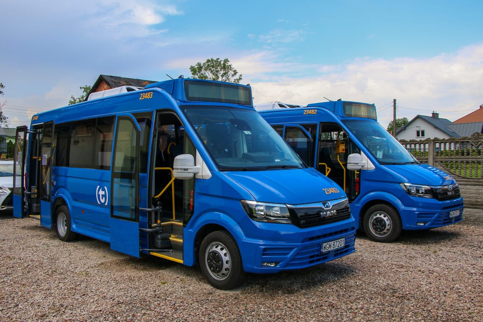 Dwa niebieskie minibusy MAN o numerach 23483 i 23482 zaparkowane na żwirowym parkingu.