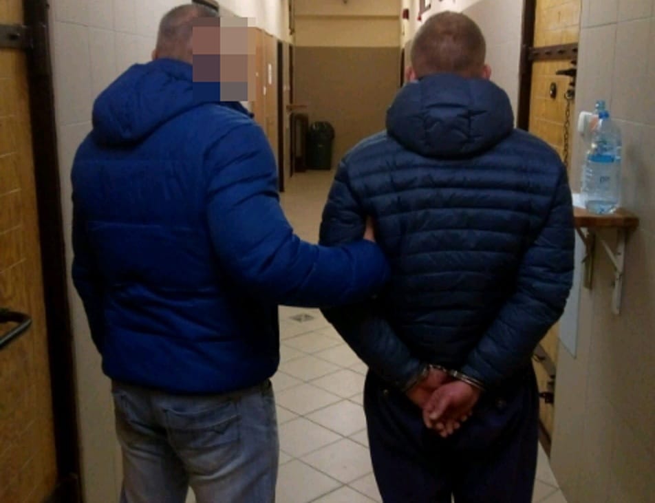 Policjant eskortujący podejrzanego w kajdankach korytarzem