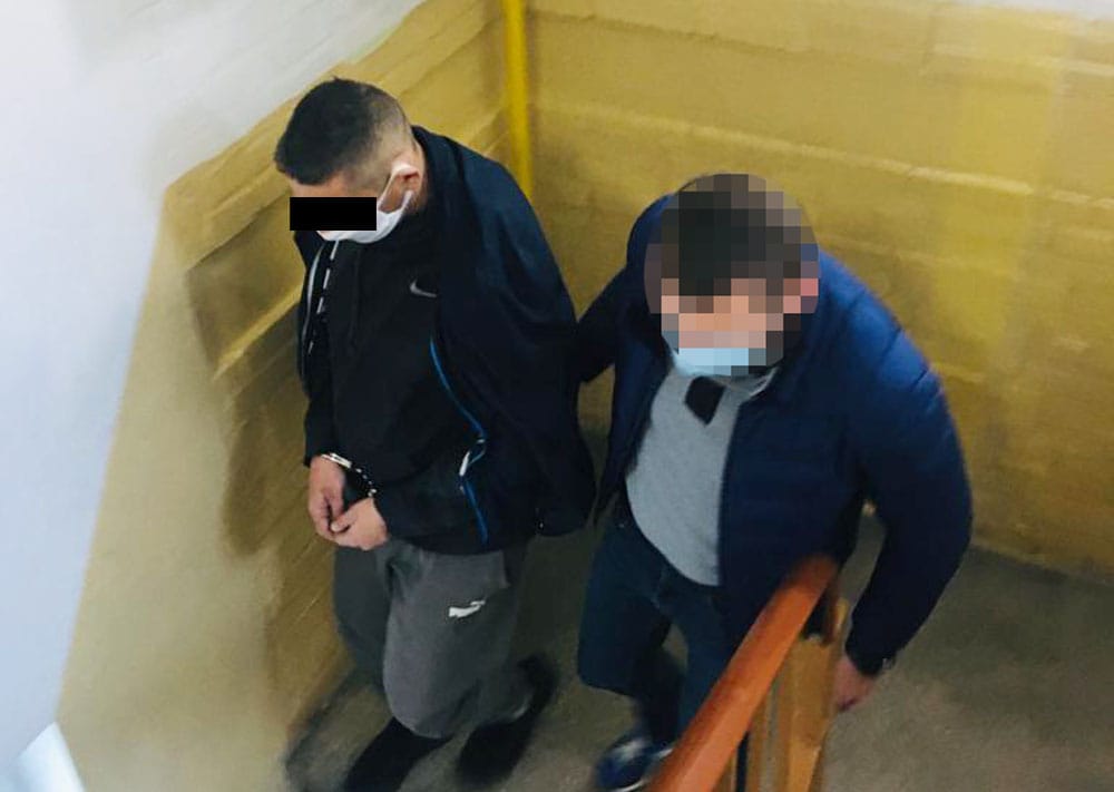 Dwóch podejrzanych w kajdankach, eskortowanych przez policję, w dół klatki schodowej.